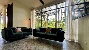 Un vaste espace de vie agrémenté de deux canapés vert foncé, d’un ventilateur de plafond et d’une grande fenêtre donnant sur un paysage verdoyant.