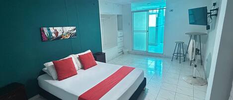 A hotel room with a bed, a desk, a chair, a TV, and a bathroom visible through a glass door.