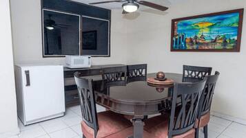 A dining area with a round table, six chairs, a ceiling fan, a white refrigerator, a microwave, and a painting on the wall.