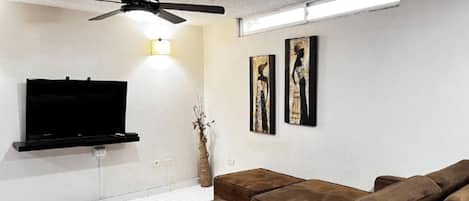 A modern living room with a flat-screen TV, a ceiling fan, a brown leather sofa, and two framed artworks on the wall.