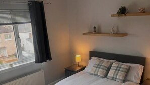 Et soveværelse med en seng, et natbord, en lampe, et vindue med gardiner og en radiator.
