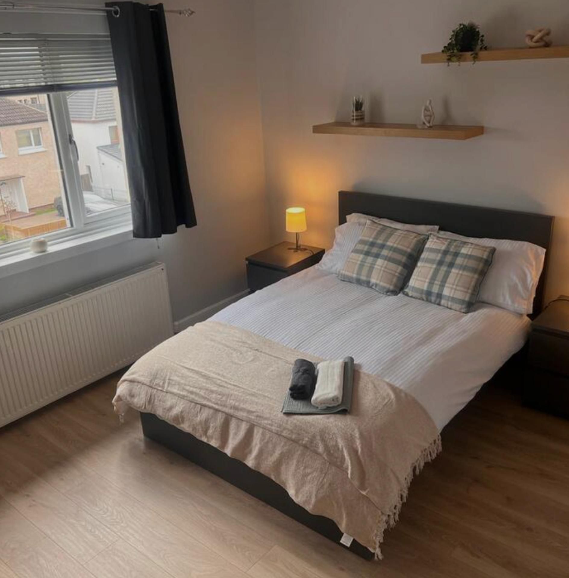 Une chambre à coucher avec un lit, une table de chevet avec une lampe, une fenêtre avec des rideaux et une étagère avec des objets décoratifs.