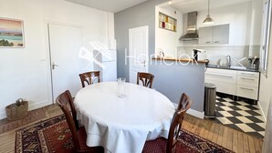 Un coin repas avec une table ronde, des chaises et un tapis à motifs. On aperçoit également une cuisine en arrière-plan, avec des placards et un évier.