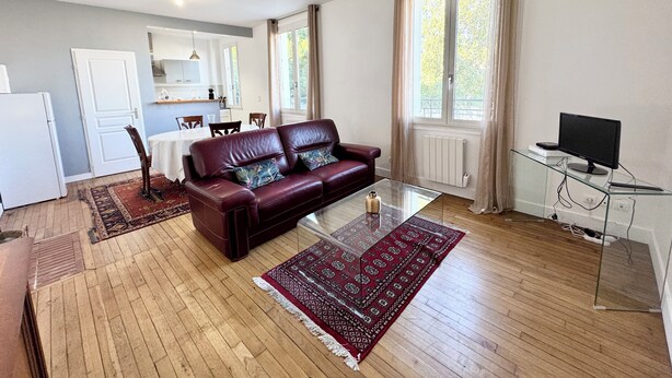 Un salon avec un canapé en cuir, une table basse en verre, un tapis à motifs rouges, un coin repas avec une table blanche et, au fond, une cuisine.