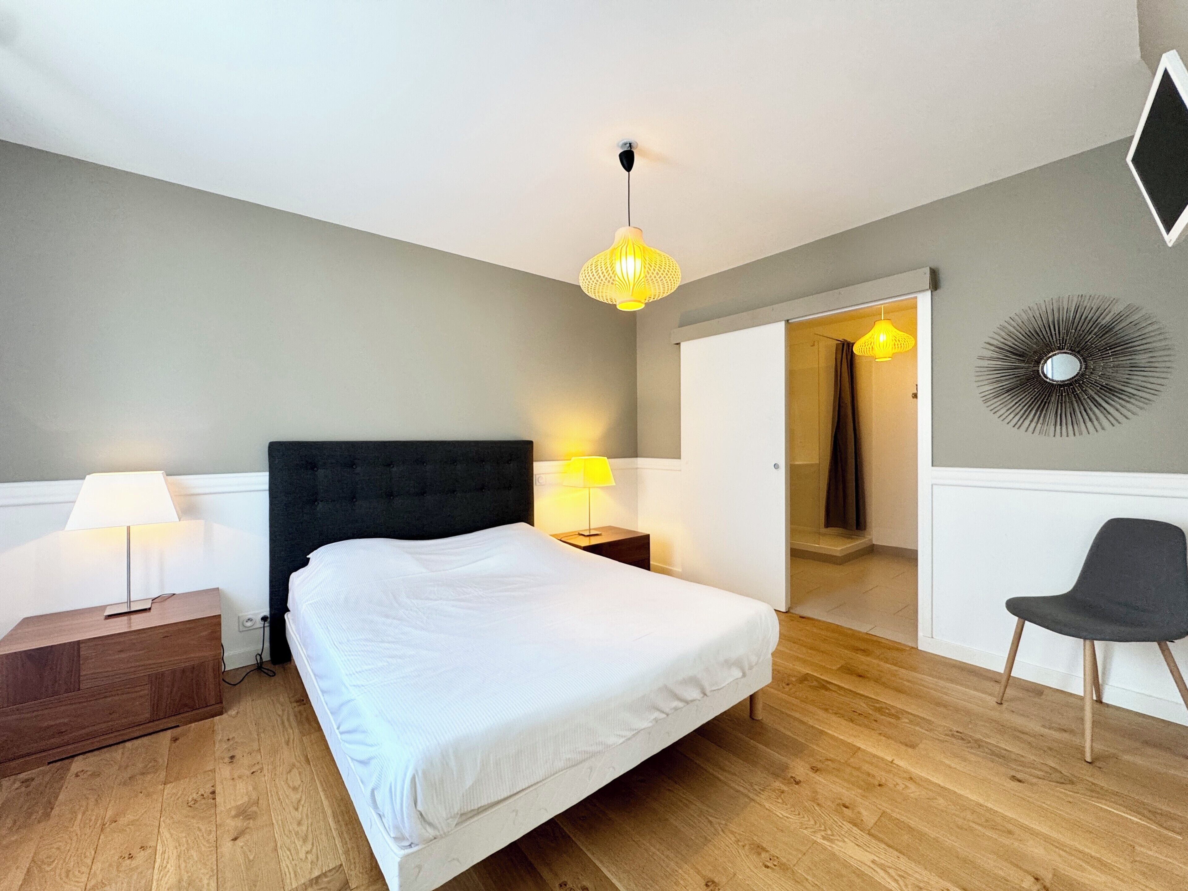 A modern bedroom with a large bed, two bedside lamps, a wooden floor, a dark headboard, and a shower area visible through an open door.