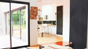 Una sala de estar moderna con un gran ventanal corredero que da a un balcón, un sofá con una manta y una cocina al fondo.