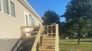 Una escalera de madera que conduce a una terraza, rodeada de grava y césped, con una casa y un árbol al fondo.