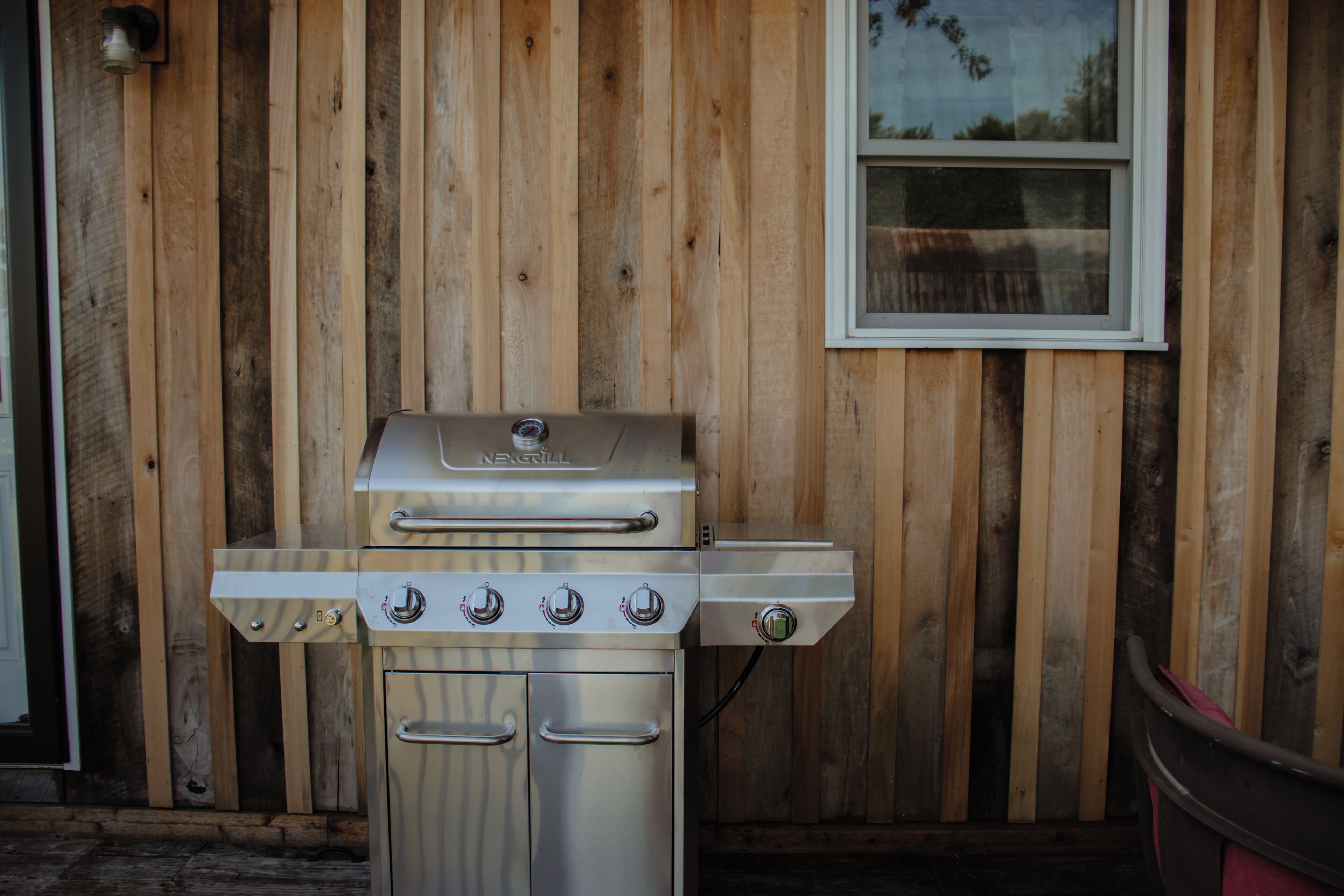 Una parrilla NERGRILL de acero inoxidable con cuatro quemadores y un quemador lateral, ubicada frente a una pared de madera con una ventana arriba.