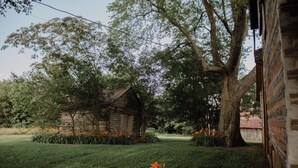 A rustic cabin with a wooden exterior, surrounded by a well-maintained lawn and flowering plants. - Rustic 1800s' Log Cabin with Modern Amenities on an Old Farm close to Owensboro (Utica)
