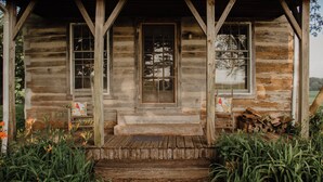 A rustic log cabin with a front porch, wooden steps, and two windows. - Rustic 1800s' Log Cabin with Modern Amenities on an Old Farm close to Owensboro (Utica)