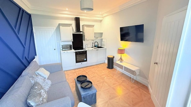 A compact living space with a kitchenette, a grey sofa, a coffee table, and a TV mounted on the wall. - Cosy Perth Apartment - City Comforts (Perth)