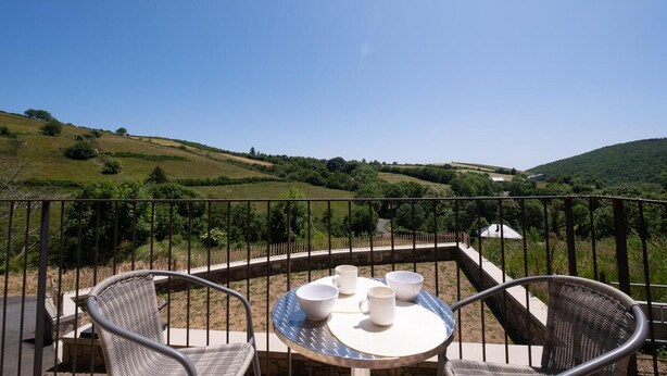 Un balcón con sillas de mimbre y una mesa, con vistas a un paisaje pintoresco con colinas y cielo despejado.