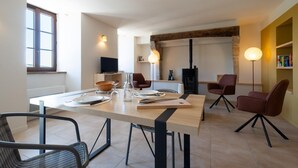 Un comedor moderno con una mesa de madera dispuesta para cuatro personas, rodeada de sillas contemporáneas. Un televisor está montado en la pared, y se observa una estantería al fondo.