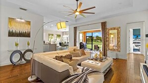 Un salon spacieux avec un grand canapé, une table basse et un ventilateur de plafond. On aperçoit un coin repas à travers une porte ouverte.