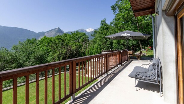 A balcony with a view of mountains, a wooden railing, a table with chairs, and an umbrella.