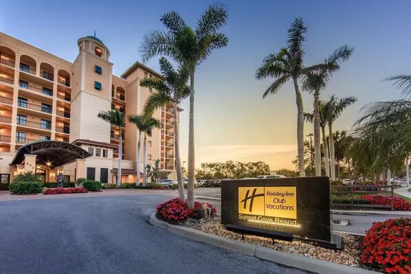 A Holiday Inn Club Vacations building with a central tower, palm trees, and a sign indicating Sunset Cove Resort.