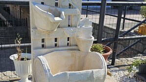 Une fontaine blanche et richement décorée, surmontée d’une statue de lion, entourée de plantes en pot et d’un lampadaire.