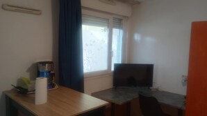 A small room with a desk, a chair, a computer, a window with curtains, and a coffee machine. - The Popes' Refuge (Avignon)