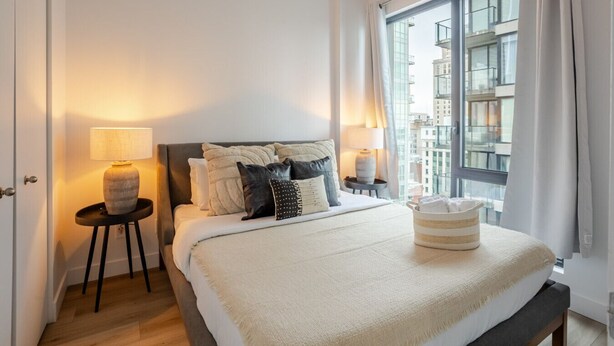 A modern bedroom with a large bed, two bedside tables, a window with a view of buildings, and a door. - Elegant 1BR Retreat | Chic Stay in Old Port (Montreal)