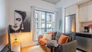 A modern living room with a sofa, a laptop, a coffee maker, and a kitchen area with a refrigerator. - Elegant 1BR Retreat | Chic Stay in Old Port (Montreal)