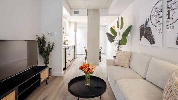 A modern living room with a flat-screen TV, a sofa, a coffee table with flowers, and a kitchen in the background.