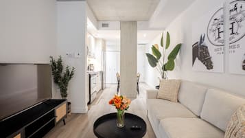 A modern living room with a flat-screen TV, a sofa, a coffee table with flowers, and a kitchen in the background.