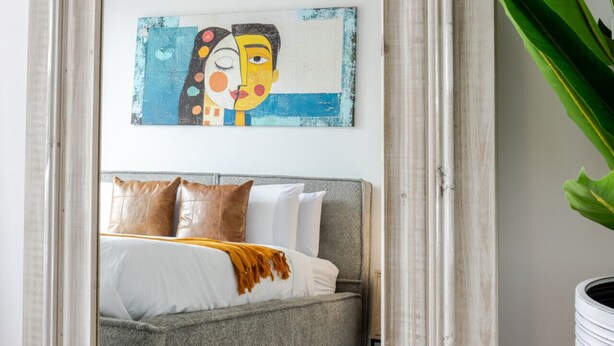 Een slaapkamer met een bed, kussens, een deken, een schilderij en een grote spiegel.