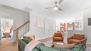 Un salon spacieux avec un canapé vert, deux fauteuils, un ventilateur de plafond et, au fond, un coin repas.