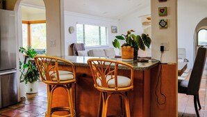 Eine Küche mit Bar, zwei Korbstühle, einer Topfpflanze und Blick zum Wohnzimmer.