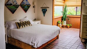 Ein Schlafzimmer mit einem hölzernen Bett, zwei an der Wand befestigten Gemälden, einem Nachttisch, einer Pendelleuchte und Blick auf einen Balkon mit Blumentöpfen.