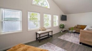 A spacious living room with large windows, a ceiling fan, a sofa, a coffee table, and a TV mounted on the wall. - Lake Front Reverie- Waneta Lake, NY (Dundee)