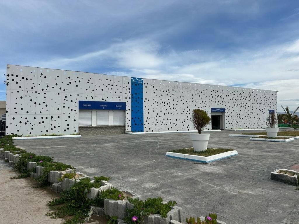 A modern building with a perforated facade, a blue vertical stripe, and a small landscaped area with a tree and plants.