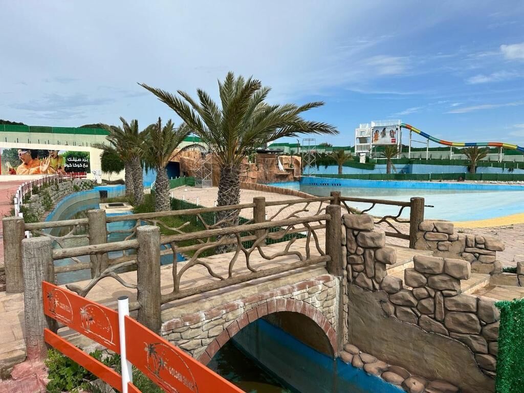 Un puente con arcos de piedra, palmeras y una zona de piscina con tobogán al fondo.
