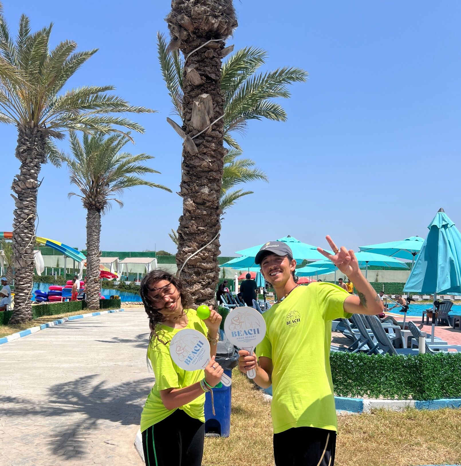Two people holding beach-themed items in a resort area with palm trees and a pool.