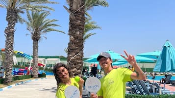 Dos personas sosteniendo objetos con temática playera en una zona turística con palmeras y una piscina.