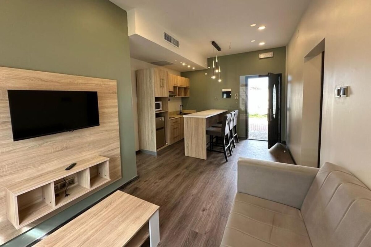 Una sala de estar moderna con televisor de pantalla plana, mueble de entretenimiento de madera, sofá color beige y una cocina con armarios de madera y una mesa de comedor.