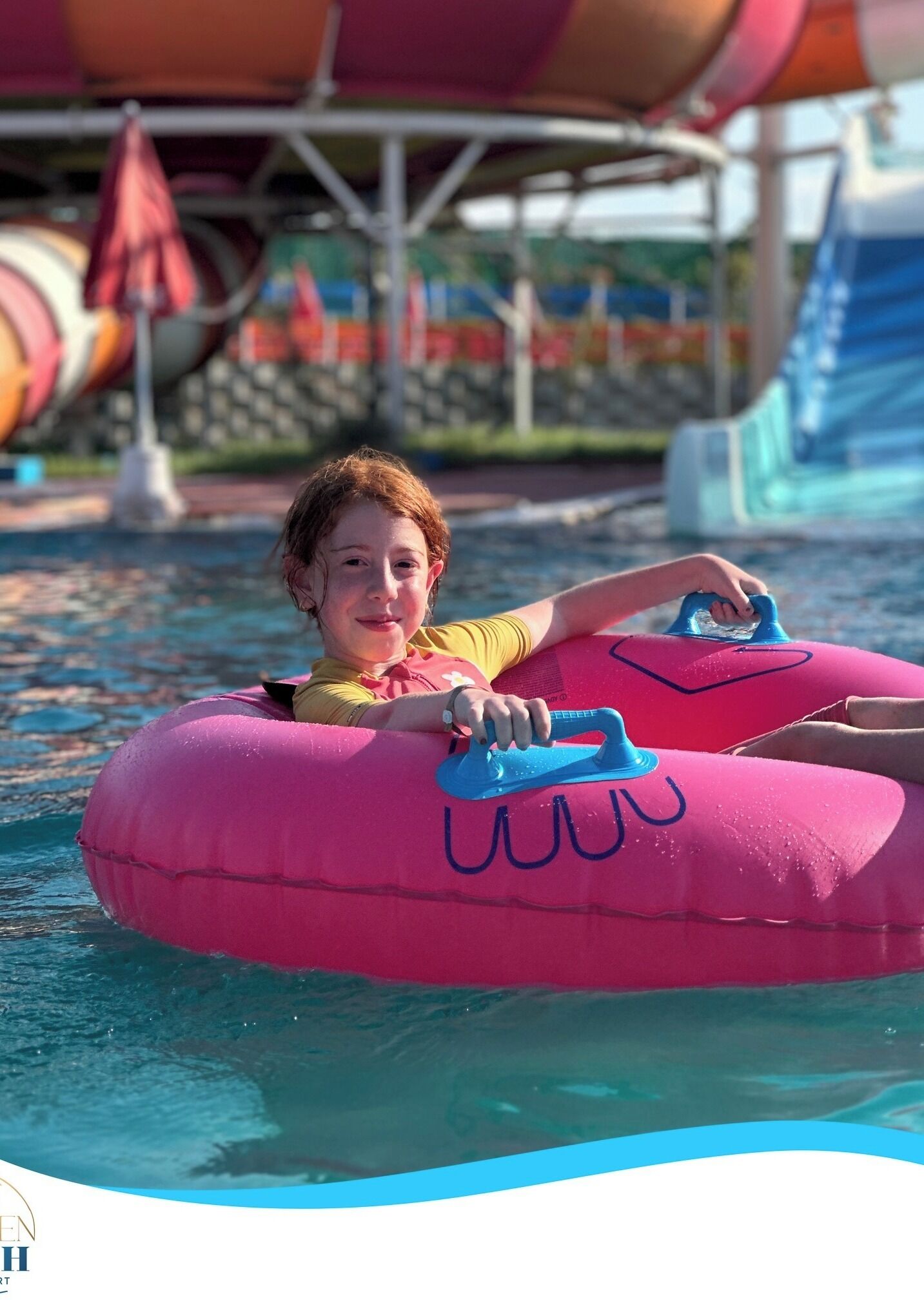 Un enfant est assis sur un bouée gonflable rose dans une zone de piscine, avec un toboggan aquatique et des parasols en arrière-plan.