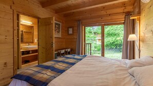 A wooden cabin bedroom with a bed, a bathroom with a sink, and a view of greenery outside.