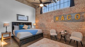 Une chambre avec un grand lit, un ventilateur de plafond, un mur en briques orné d’une enseigne « WACO » et une petite table avec des chaises.