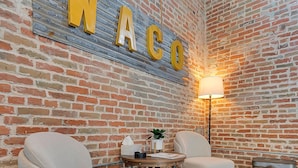 Une pièce chaleureuse avec un mur en briques, deux fauteuils rembourrés, une petite table avec une lampe et un panneau décoratif sur lequel on peut lire le mot « WACO ».