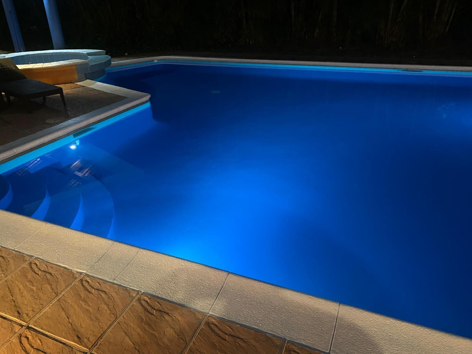 Une piscine avec un bord carrelé et un espace de détente comprenant une chaise et une table.