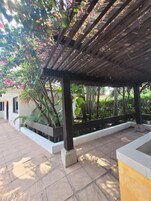 A covered outdoor area with a pergola, tiled flooring, and lush greenery including flowering plants.
