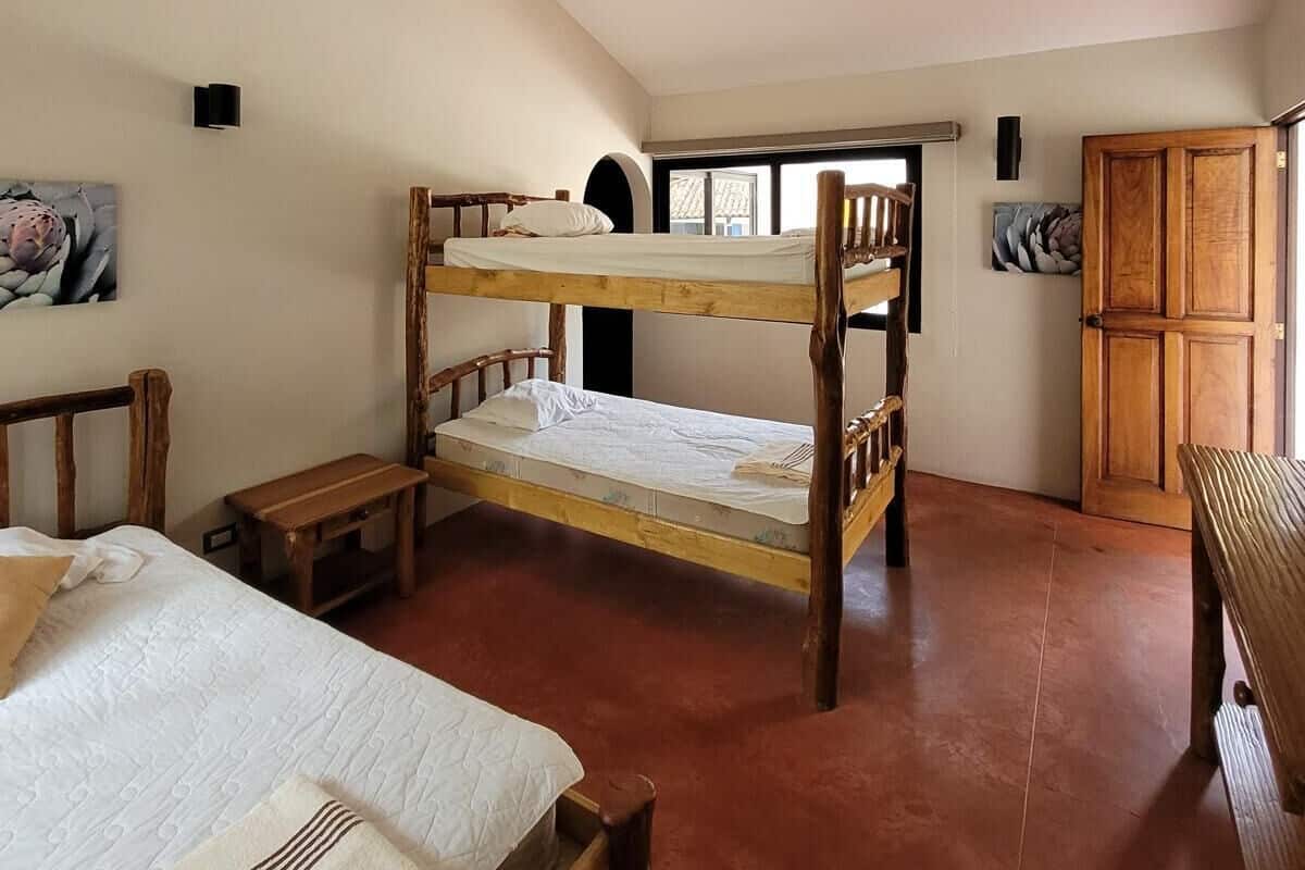 Ein Schlafraum mit Etagenbetten, einem hölzernen Kopfteil, einem kleinen Holztisch und einer Bank. Es gibt einen Kleiderschrank und ein Fenster mit Blick nach außen.