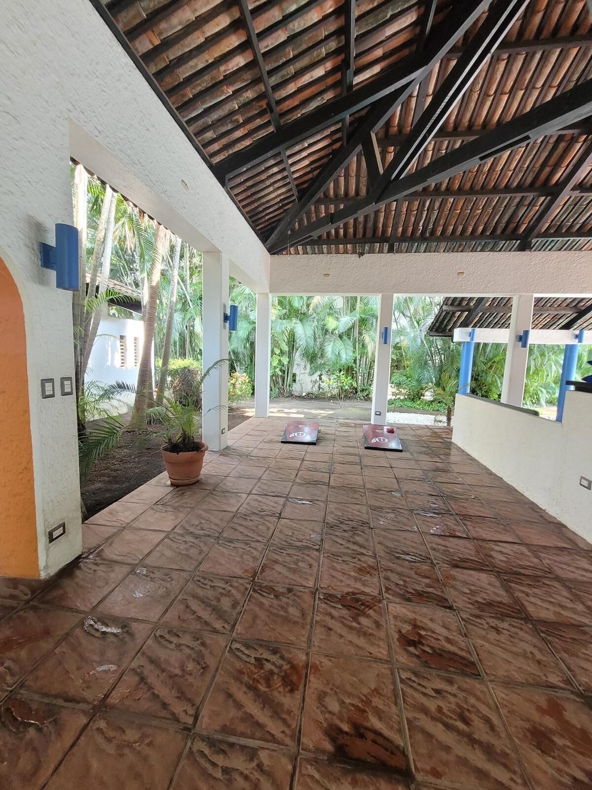 A covered outdoor area with tiled flooring, potted plants, and a view of greenery.