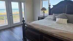 Ein Schlafzimmer mit einem großen Bett, einem Nachttisch und einer Lampe. Durch eine Glastür ist der Blick auf den Strand möglich.
