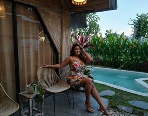A person sitting on a wicker chair by a poolside, with a potted plant and a small table nearby. - Rustic 2 bedrooms villa near Obsidian Gym (Bali)
