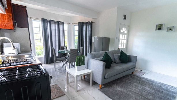 A modern living room with a grey sofa, a dining table with chairs, a kitchen area with a stove, and a large window with curtains.