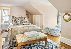 A bedroom with a bed, a bench, a round ottoman, a mirror, and a window with curtains.