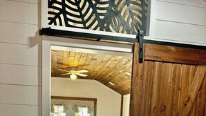 A room with a wooden ceiling, a bed, a sofa, and a view through a doorway to another room with a window and a door. - Mini Mallard at Reelfoot Lake (Hornbeak)