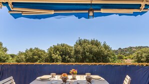 Zona pranzo all'aperto con un tavolo imbandito per due, circondato da sedie, sotto un tendone blu.